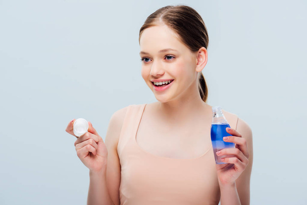happy smiling teenage girl holding cosmetic lotion and looking away isolated on grey - Photo, Image