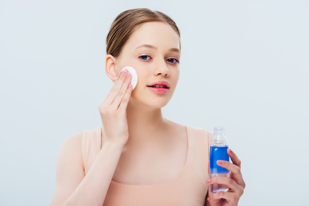 pretty teenage girl applying lotion on face with cotton pad isolated on grey - Foto, Imagen