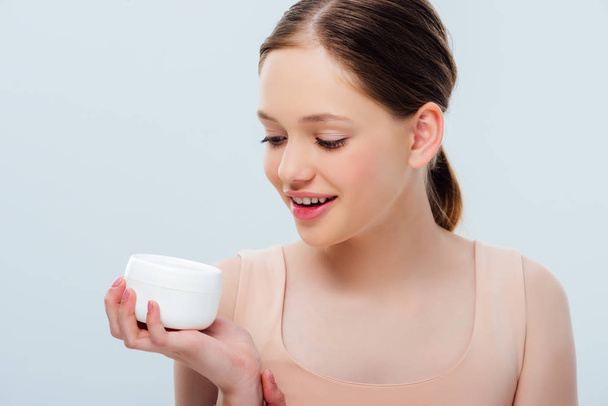 cheerful teenage girl looking at cosmetic cream isolated on grey - Foto, Imagem