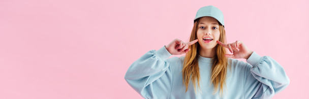 panoramic shot of pretty teenage girl in cap pointing with fingers at smile isolated on pink - Foto, immagini