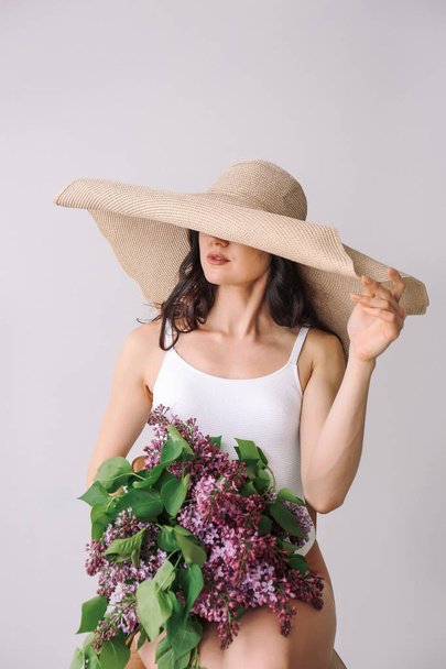 Hermosa joven en traje de baño y con ramo de flores lila sobre fondo blanco
 - Foto, Imagen
