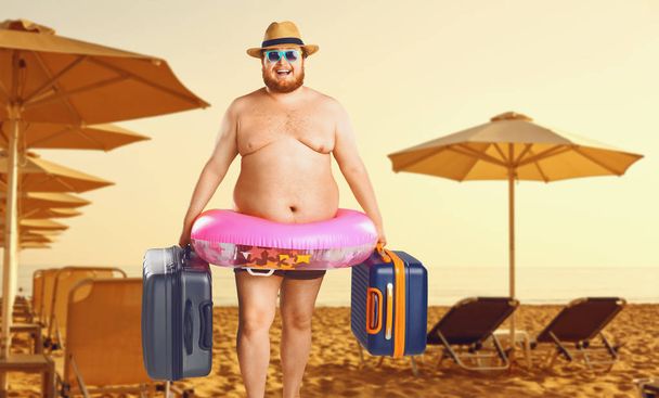 夏のビーチを背景にスーツケースとゴムリングを持つ水着を着た太い男. - 写真・画像