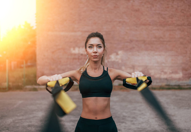 Female athlete training fitness workout in summer city. Rubber loop strap, push-up wiring hands. Active lifestyle, sportswear leggings top. Emotions concentration strength of the power exercises. - Photo, image