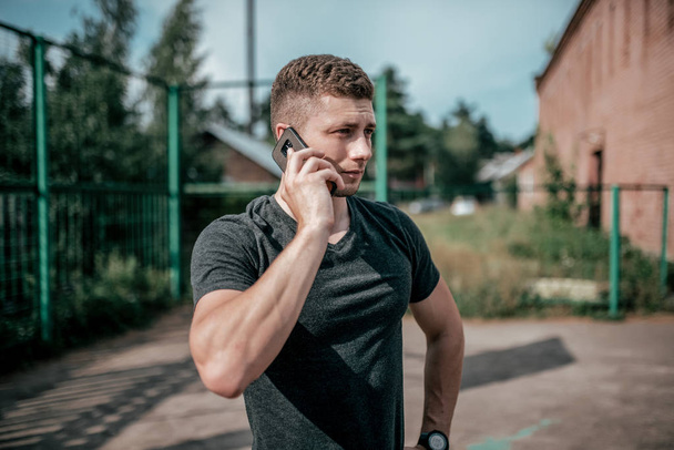 A man athlete calls on the phone, after a workout, rest after a fitness workout jog. Summer in the city on the background of the athletic field. Concept of online application call in social networks. - Valokuva, kuva