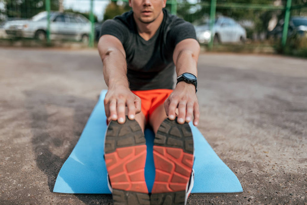 A man athlete doing workout on mat, gymnastics, training, fitness workout flexibility. Summer city on background athletic field. Active lifestyle youth, close-up hand sneakers smart bracelet watches. - Фото, зображення