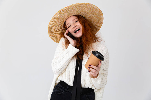 Young cute girl redhead posing isolated over white wall background drinking coffee talking by phone. - Valokuva, kuva