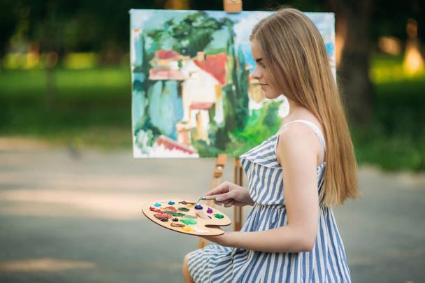 Prachtig blond haar meisje zit op kruk en tekent u een afbeelding in het park met behulp van een palet met verf en een spatel - Foto, afbeelding