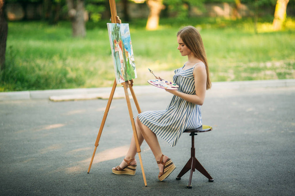 Hermosa chica de pelo rubio sentada en el taburete y dibuja un cuadro en el parque usando una paleta con pinturas y una espátula
 - Foto, Imagen