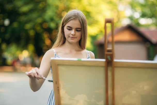 Güzel kız parkta boyalar ve bir spatula ile bir palet kullanarak bir resim çizer. Şövale ve tuval resmi - Fotoğraf, Görsel