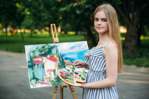 Hermosa artista chica dibuja un cuadro en el parque utilizando una paleta con pinturas y una espátula. caballete y lienzo con una imagen
 - Foto, Imagen