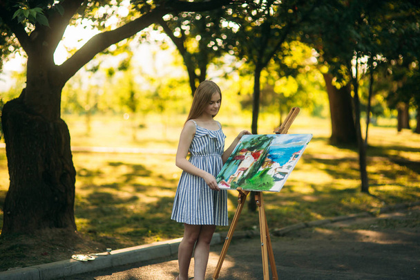 Beautiful girl artist draws a picture in the park using a palette with paints and a spatula. Easel and canvas with a picture - Photo, Image
