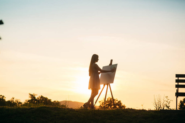 Silhouet van een blond meisje schildert een schilderij op het canvas met de hulp van verven. Een houten ezel houdt de afbeelding. De zomer is een zonnige dag, zonsondergang - Foto, afbeelding