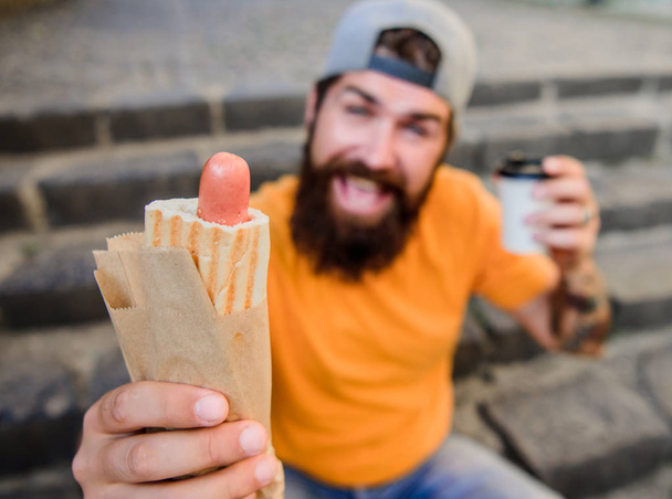 Urban lifestyle nutrition. Carefree hipster eat junk food while sit on stairs. Hungry man snack. Junk food. Guy eating hot dog. Man bearded bite tasty sausage and drink paper cup. Street food so good - Photo, Image