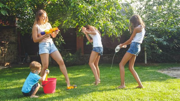 Photo of happy laughing family splashing water with water guns and garden hose at backyard. People playing and having fun on hot sunny summer day - Photo, Image