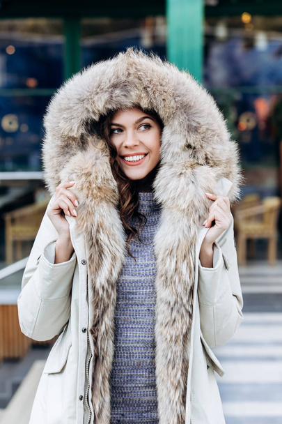 Attractive stylish girl dressed in a knitted dress and a light coat with fur poses in the street - Foto, Imagen