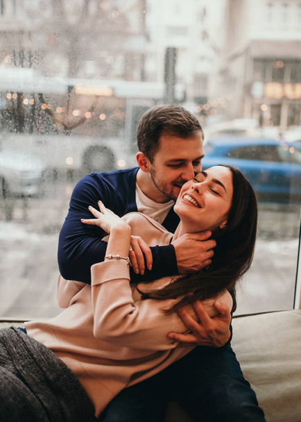 Loving guy hugs his beautiful girlfriend sitting on the windowsill in a cozy cafe. - Photo, Image