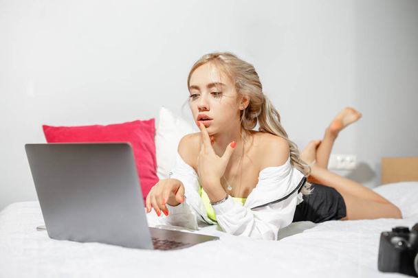 Young girl blogger dressed in fashionable clothes lying on the bed next to the camera and working at the laptop in the room - 写真・画像