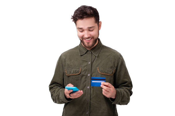 Gelukkig glimlachende jonge kerel met behulp van een mobiele telefoon en creditcard over witte muur  - Foto, afbeelding