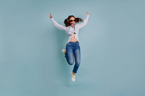 Podekscytowany Europejska Urocza dziewczyna z latającymi włosami noszących dżinsy i skoki koszuli i ma zabawę na białym tle niebieskim  - Zdjęcie, obraz