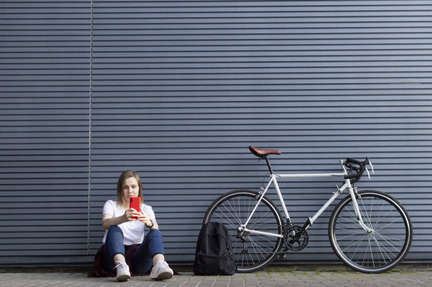 beautiful young girl with a smartphone, a backpack and a bicycle sits on the background of the wall, she stopped to rest - 写真・画像