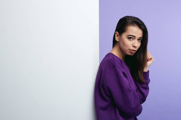Pretty young girl, standing alongside empty white billboard wall isolated over purple background. - Foto, Imagem