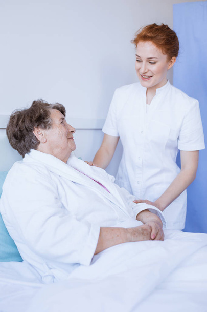 Старша сіра жінка лежить у ліжку білої лікарні з молодою корисною медсестрою, тримаючи її за руку
 - Фото, зображення