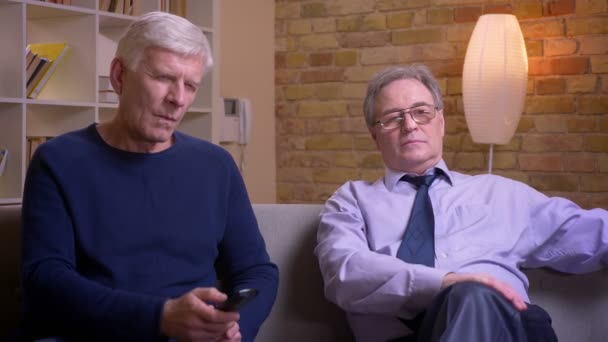 Portrait of senior male friends sitting together on sofa watching TV and discussing being attentive and serious. - Video