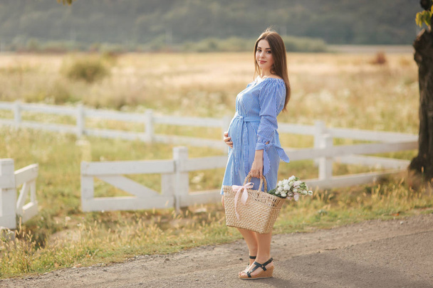 Belle femme enceinte en robe bleue marchant entendre la ferme. Sac tricoté avec bouquet de fleurs. Contexte de l'arbre et des montagnes
 - Photo, image