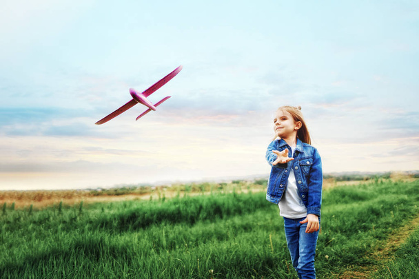 Little girl, launches a toy plane, into the air against, the background of green grass. Child launches a toy plane. Beautiful little girl, stands on the grass and launches a pink toy plane - Foto, Imagen