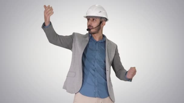 Architect man in helmet and head set talking to camera and making gestures showing on some objects on gradient background. - Imágenes, Vídeo