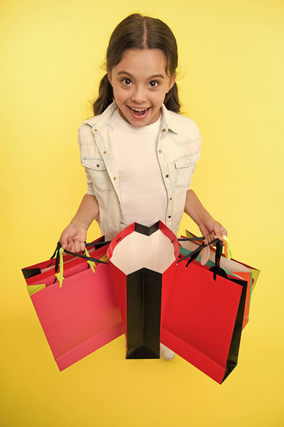 Check out her profitable purchases. Girl carries shopping bags yellow background. Girl fond of shopping. Child cute shopaholic with bunch shopping bags black friday total sale. Exciting shopping - Fotó, kép