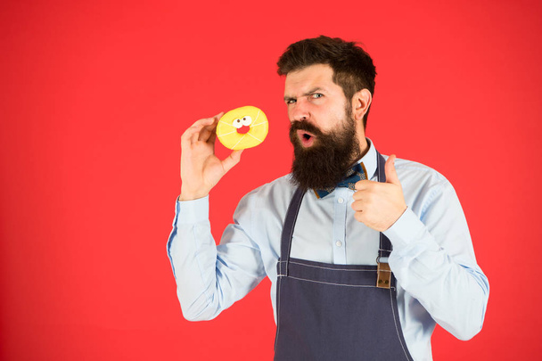 Hipster bearded baker hold glazed donut on red background. Cafe and bakery concept. Sweet donut from baker. Man bearded baker in apron hold cute dessert. Private bakery business. Just try this - Фото, изображение