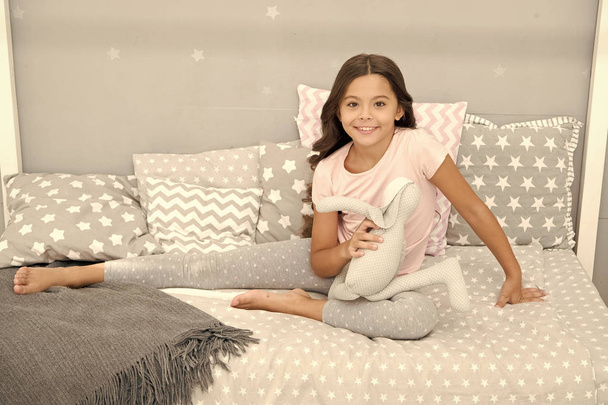 Girl long curly hair enjoy evening time with favorite toy. Kid sit bed and play bunny toy modern bedroom interior. Evening time. Girl child wear pajamas play bunny toy. Play soft toy before go sleep - Foto, immagini