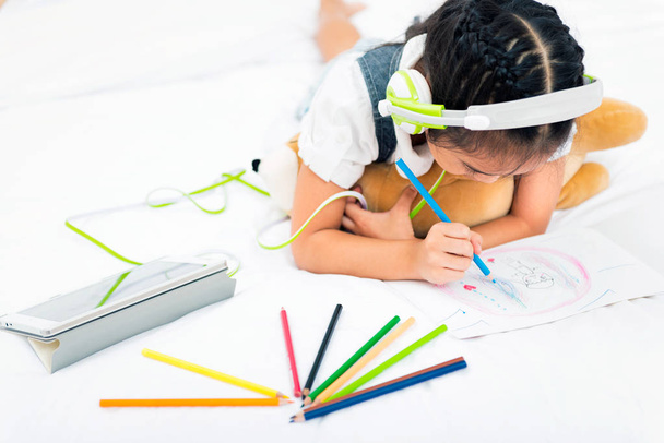 Азіатські дівчинка слухати музику з планшета з зеленими навушник під час написання з барвистими олівці на ліжку в будинку. Освіта для дітей і зміцнює уяву дітей концепція. - Фото, зображення