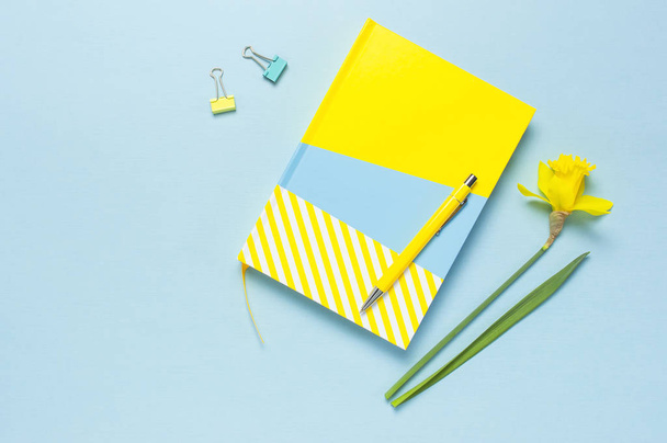 Yellow-blue notebook, pen, clips, spring flowers daffodils narcissus on blue background. Female desktop, Office desk, spring concept. Flat lay, top view, copy space. Template for feminine blog - Photo, image