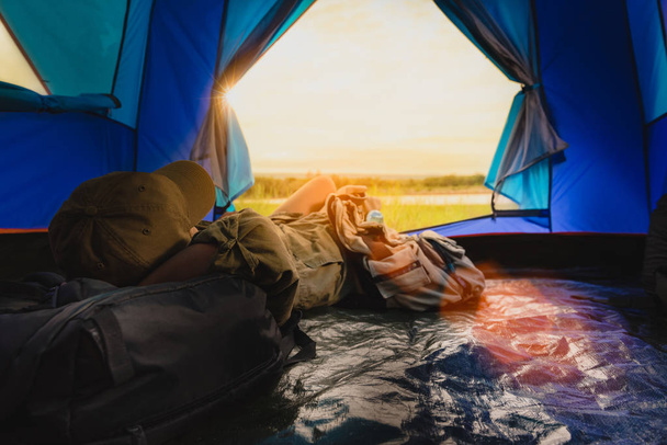 Mujer joven acostada en tienda de campaña o durmiendo en el bolso con una vista al atardecer en las montañas y el lago, Camping con puesta de sol en el campamento base, Una actividad Relajante en verano
 - Foto, Imagen