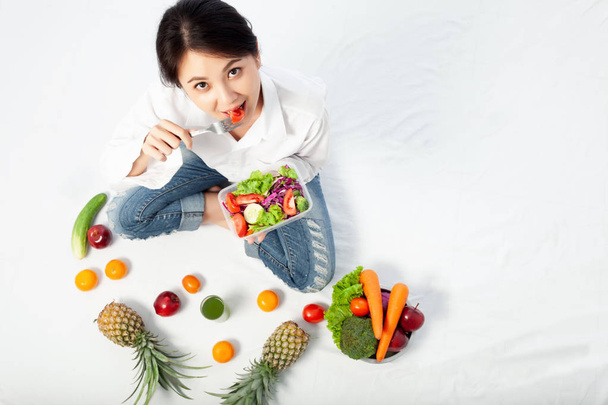 Вид сверху на азиатскую молодую женщину, счастливую есть здоровый салат и сидеть со свежими фруктами и здоровой диетой в белой комнате
. - Фото, изображение