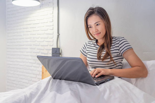 Nuori aasialainen nainen leikkii laptop hänen sängyllä valkoinen bedroo
 - Valokuva, kuva