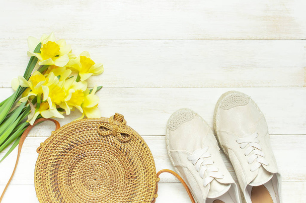 Fashionable natural organic round rattan bag, beige women's espadrilles yellow narcissus daffodil flowers on light wooden background flat lay Trendy bamboo bag Ecobags from Bali Summer fashion concept - Photo, Image