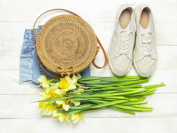 Fashionable natural organic round rattan bag, denim shorts, beige women's espadrilles, yellow narcissus daffodil flowers on light wooden background flat lay. Summer fashion concept. Ecobags from Bali - Photo, Image