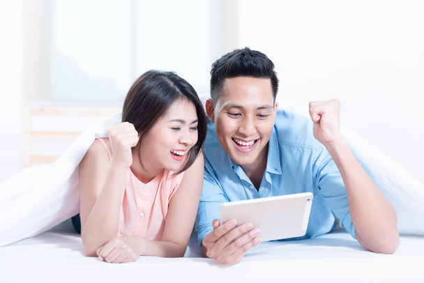 asiatisches junges Paar mit einer Tablette, während es auf dem Bett im Schlafzimmer liegt. High-Speed-Internet 5g Service-Technologie zu Hause, Telekommunikationsnetze entworfenes Konzept. - Foto, Bild
