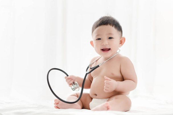 Asiático bebé niño sonriendo y juega estetoscopio en la cama un fondo blanco. Salud para bebés y habilidades desarrollan concepto
. - Foto, imagen