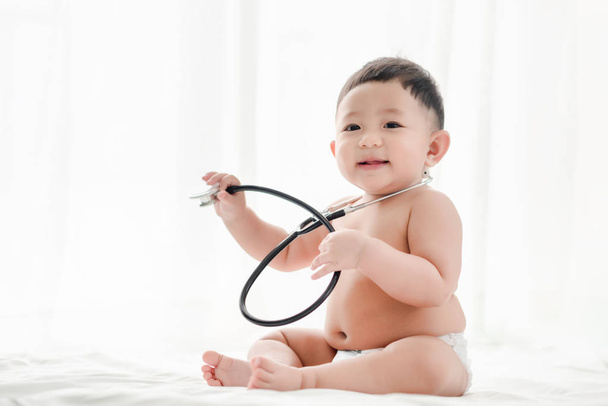 Ασιατικό μωρό αγόρι χαμογελά και παίζει στηθοσκόπιο στο κρεβάτι ένα λευκό φόντο. Υγειονομική περίθαλψη για το μωρό και την ανάπτυξη δεξιοτήτων. - Φωτογραφία, εικόνα