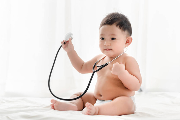 Asya erkek bebek gülümseyen ve yatakta beyaz bir arka plan üzerinde stetoskop oynar. Bebek için sağlık ve beceri kavramı geliştirmek. - Fotoğraf, Görsel
