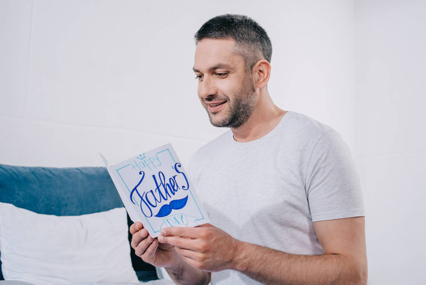 feliz hombre sosteniendo padres día tarjeta de felicitación hecha a mano mientras está sentado en el dormitorio
 - Foto, Imagen