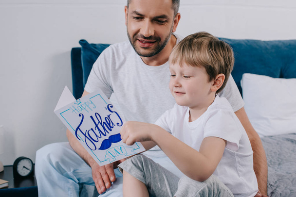 smiling kid holding fathers day greeting card while sitting near happy father - Fotoğraf, Görsel