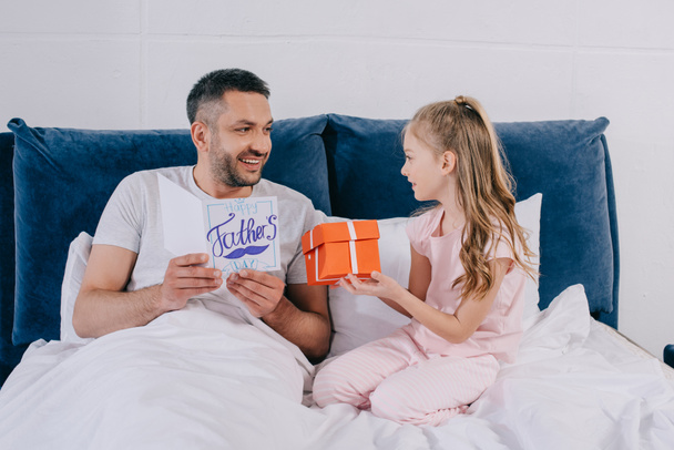 happy man holding fathers day greeting card while sitting near daughter holding gift box - Foto, Imagem