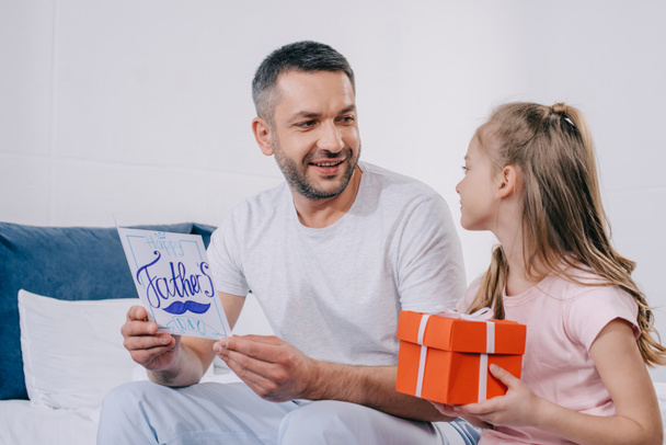 adorable daughter sitting with gift box near happy dad holding fathers day greeting card - Foto, afbeelding