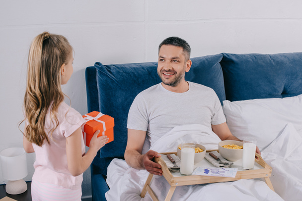 adorable daughter holding fathers day gift box while standing near father having breakfast in bed - Φωτογραφία, εικόνα