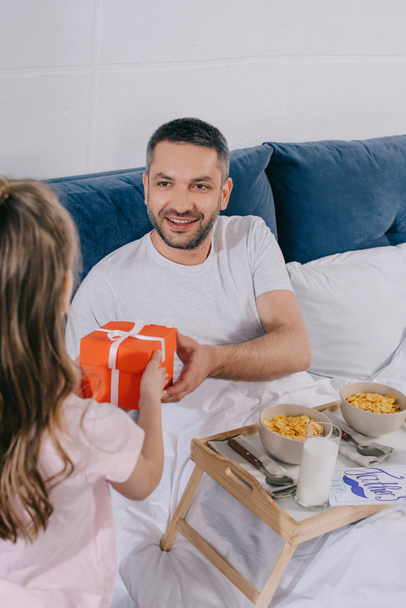 back view of cute child presenting fathers day gift box to smiling father having breakfast in bed - Φωτογραφία, εικόνα